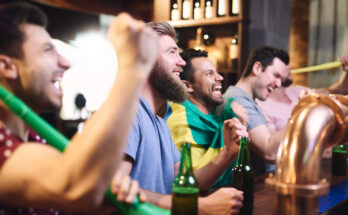 A imagem é a foto de quatro homens, que estão em balcão de bar e assistindo a um jogo de futebol.