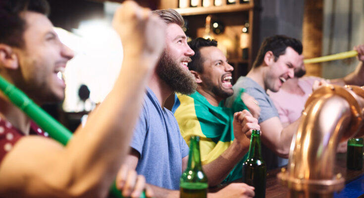 A imagem é a foto de quatro homens, que estão em balcão de bar e assistindo a um jogo de futebol.