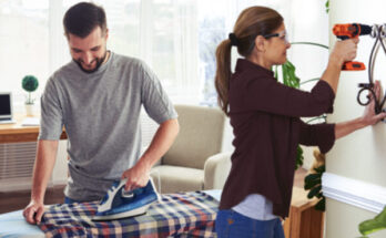 A imagem é a foto de casal, homem e mulher, ambos os dois estão utilizando produtos da marca Black & Decker. Ele está com ferro de passar roupa e ela, com furadeira.