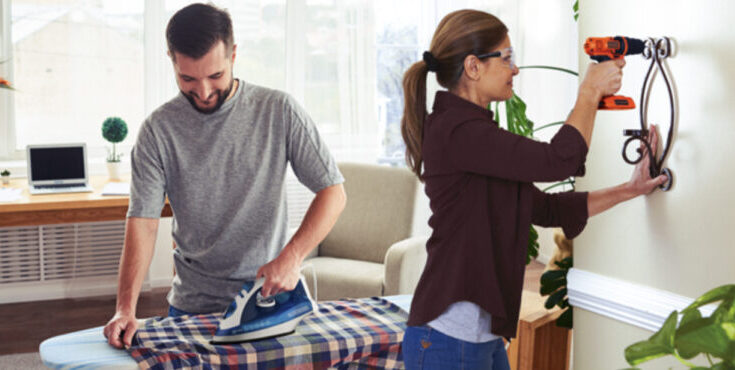 A imagem é a foto de casal, homem e mulher, ambos os dois estão utilizando produtos da marca Black & Decker. Ele está com ferro de passar roupa e ela, com furadeira.